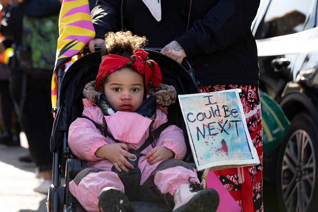 A child sits in a stroller with a sign that reads “I could be next.”