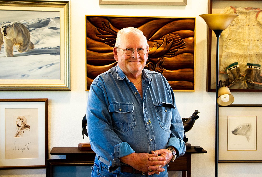 Doug Hudson stands in front of his collection of artwork, accumulated over many years. Directly behind him hangs one of his numerous wood carvings, inspired by his Alaskan roots.